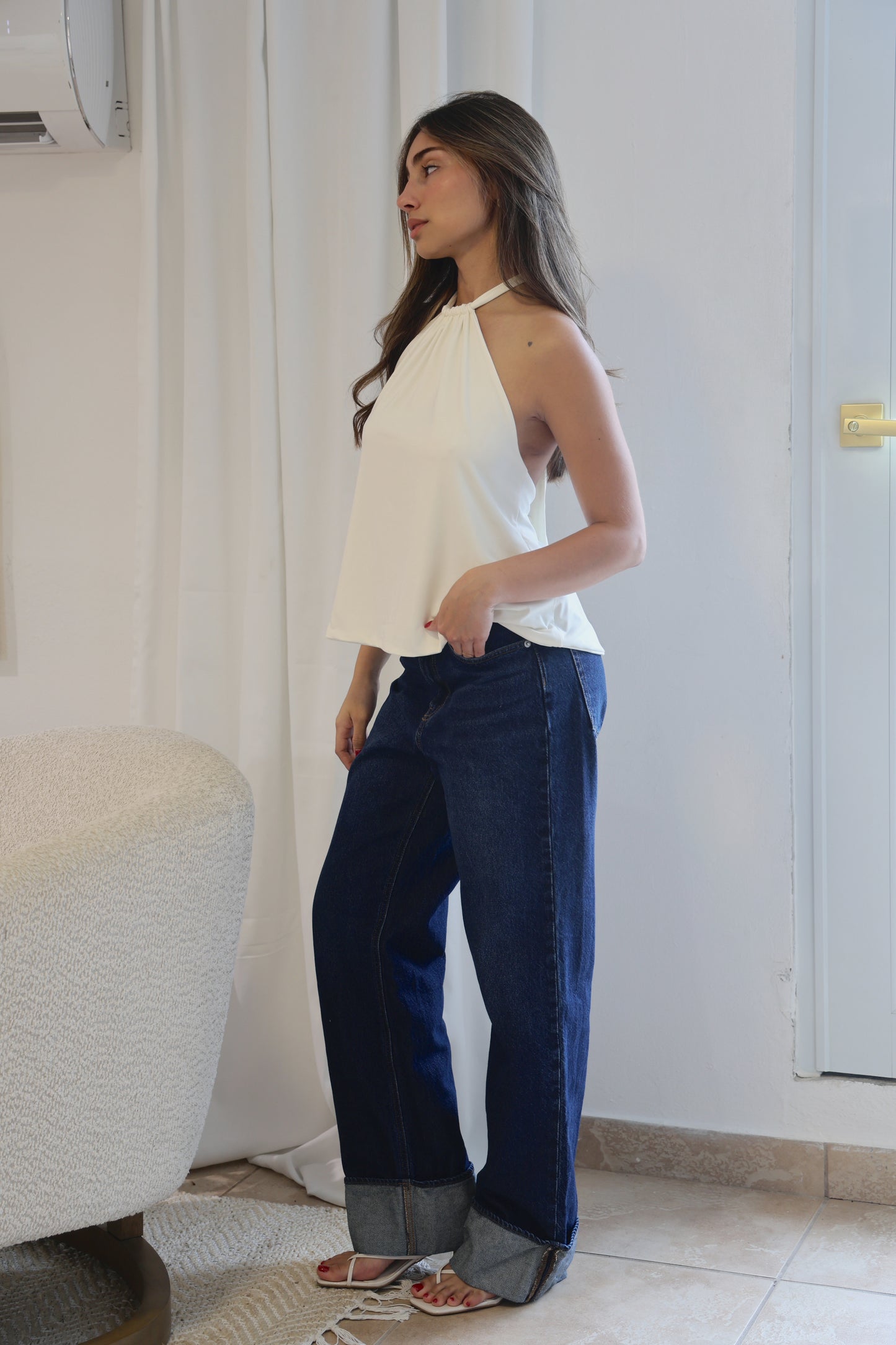 Woman wearing a white top and blue pants standing in a room with a white curtain and tiled floor.