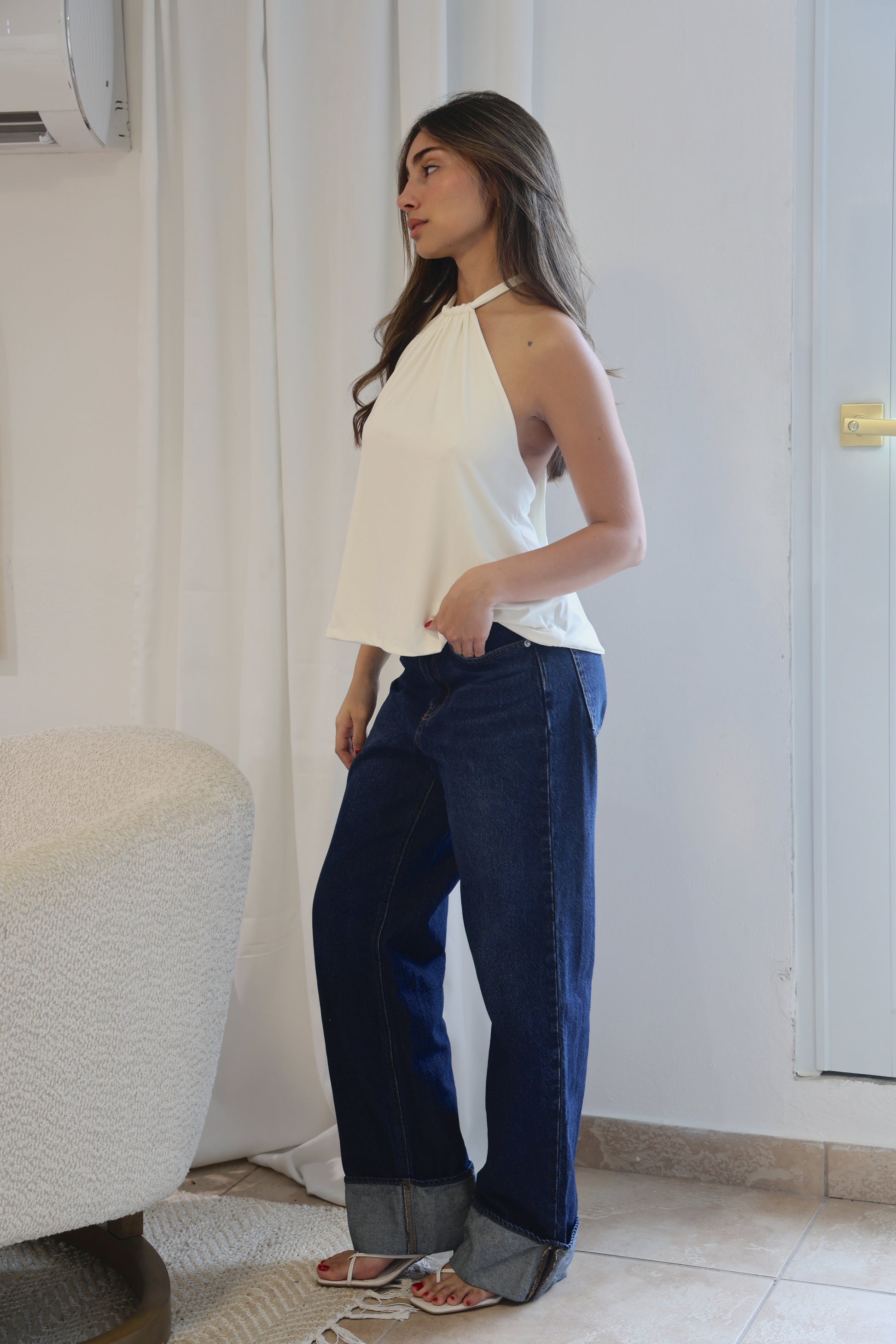 Woman wearing a white top and blue pants standing in a room with a white curtain and tiled floor.